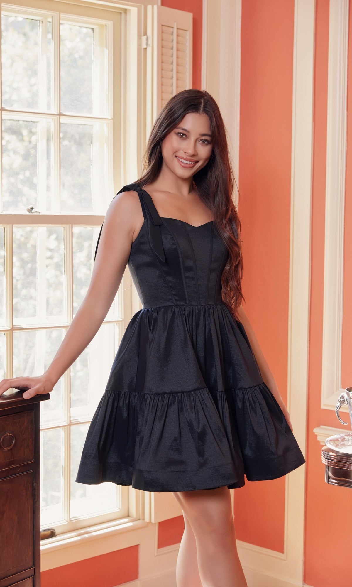 A woman stands indoors by a window, smiling at the camera in the Shoulder-Tie Short Lace-Up A-Line Hoco Dress T869—a stylish sleeveless black dress with a fitted bodice and flared skirt, perfect for any semi-formal party.